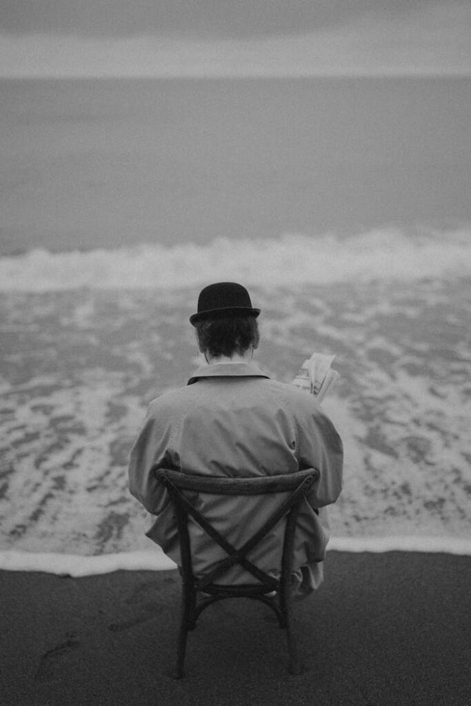 A man in a hat reads a newspaper by the sea, evoking a sense of solitude and reflection.